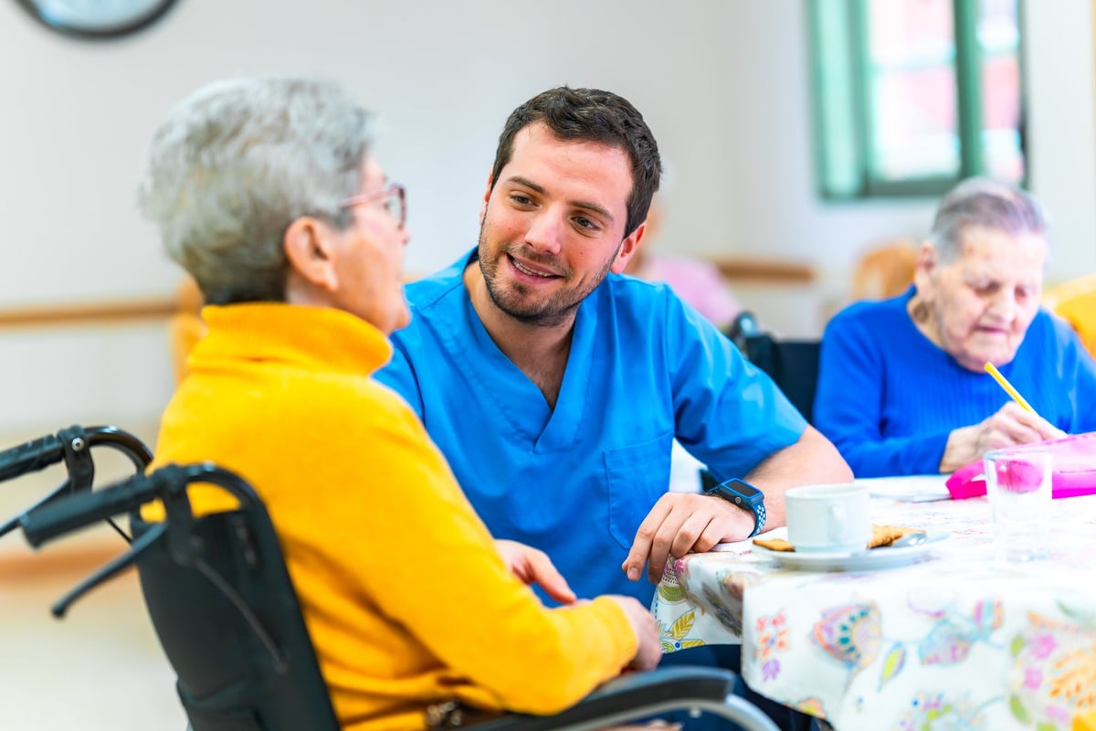 Male nurse providing care and support to an elderly woman in a wheelchair at a nursing home, engaging in conversation and ensuring her well-being