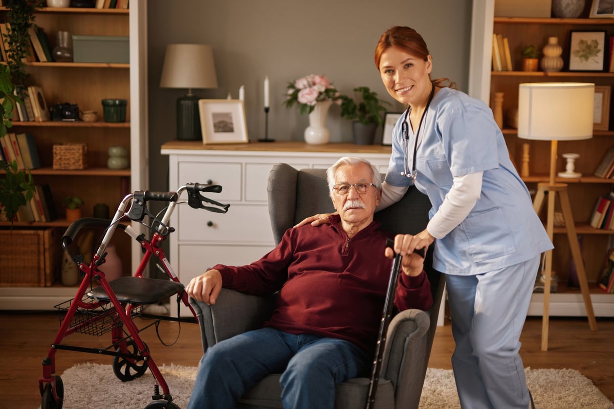 Nurse smiling and assisting elderly man at home with walking cane