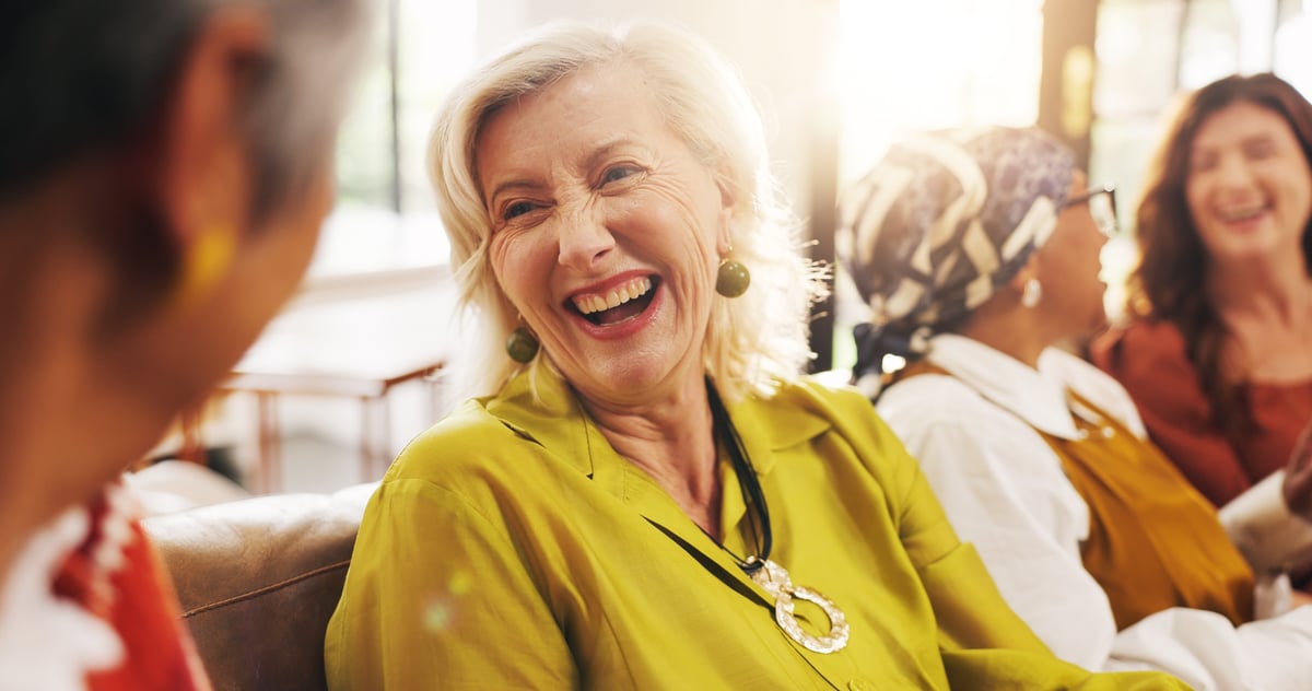 Laughing senior women bonding with friends on a couch in a home lounge, happy and social
