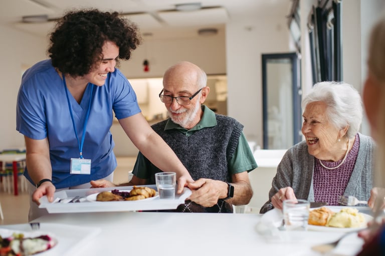 Caregiver serving lunch to elderly residents in a care home dining room with a diverse group of seniors